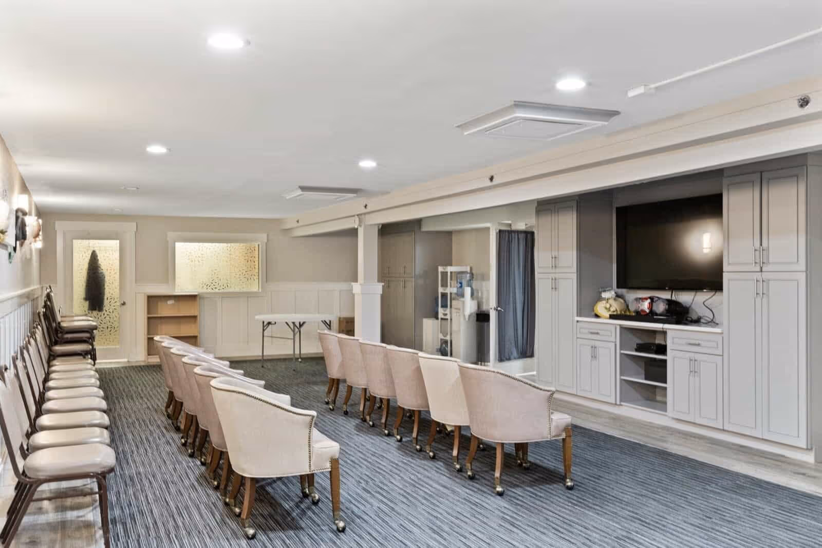 A spacious common room with two rows of chairs facing a large flat-screen TV mounted on a wall with built-in gray cabinetry. The room has carpeted flooring, recessed ceiling lights, and a water cooler in the corner. There are additional chairs lined up against the left wall and a folding table near the back.