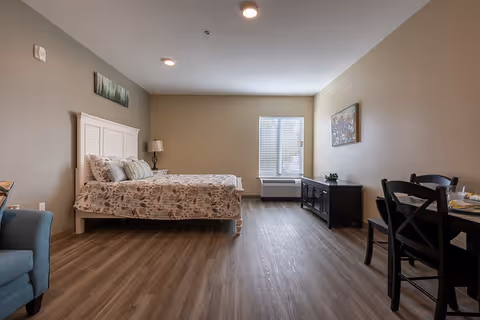 Studio bedroom with a bed, nightstand and lamp, armchair, dining table, and TV console beneath a window.