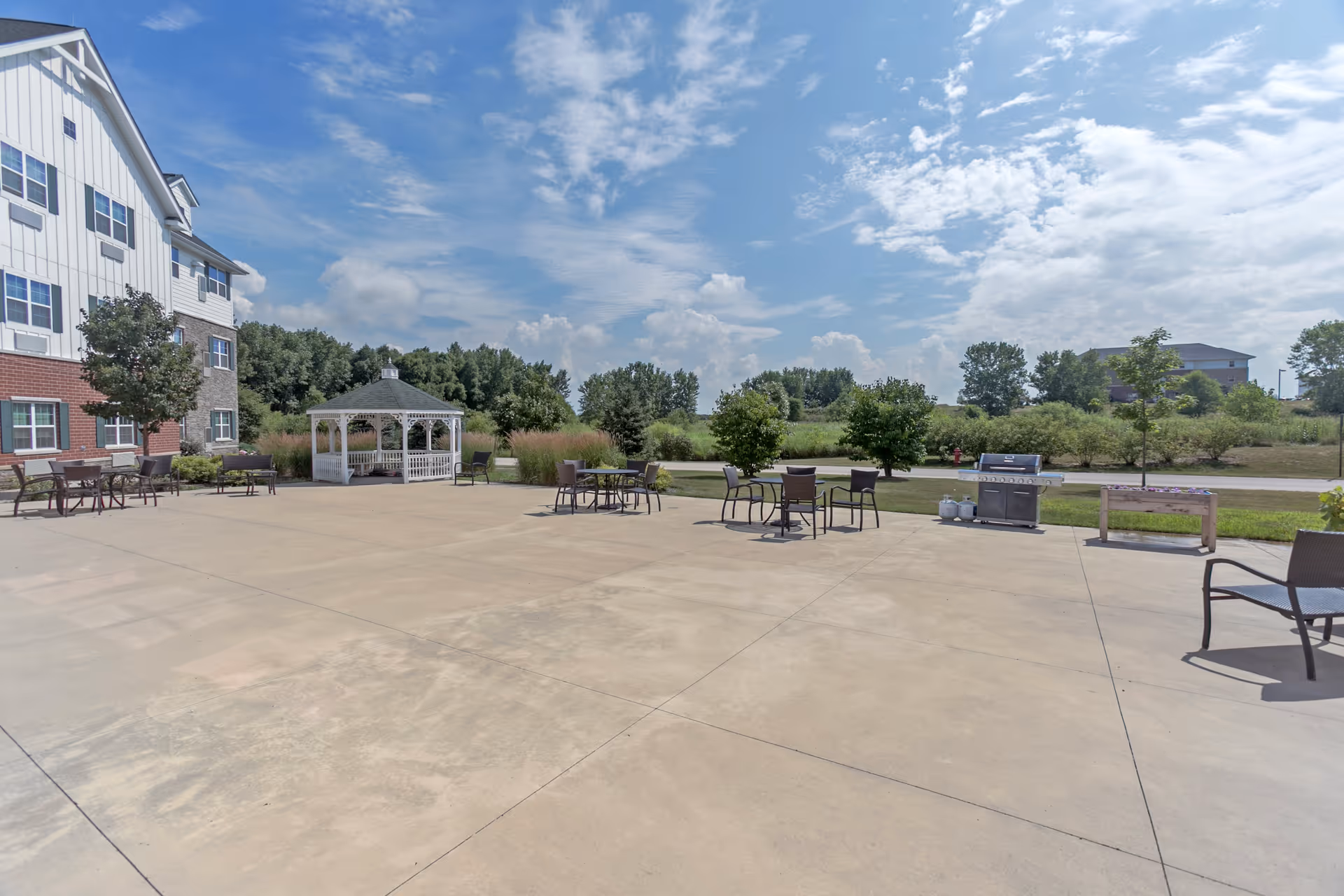 Outdoor patio area at Deer Path of Huntley with several tables and chairs, a white gazebo, a barbecue grill, and a planter box. The patio is adjacent to a multi-story building with a mix of brick and siding exterior. Trees and greenery surround the area under a partly cloudy blue sky.