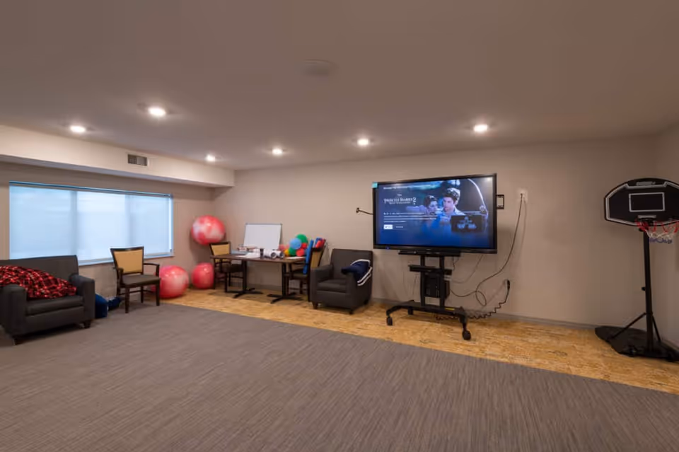 A communal activity room with chairs, a table, exercise balls, a large flat-screen TV on a rolling stand, and a small basketball hoop.