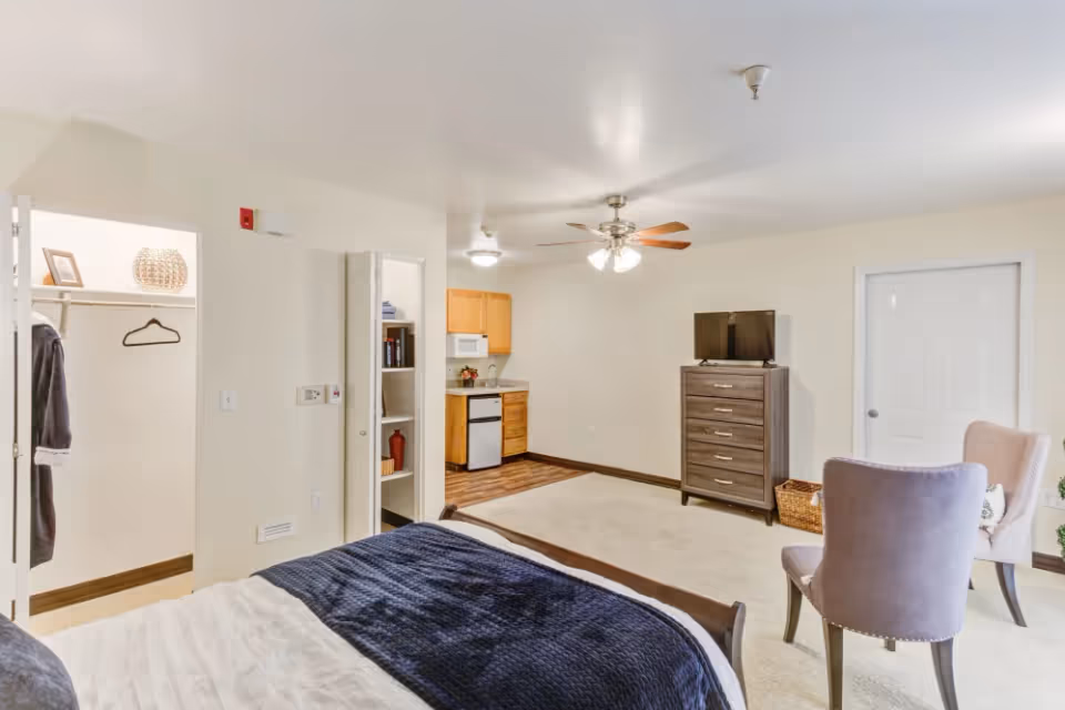 A senior living facility room featuring a bed with a dark blue blanket, an open closet with a robe and hanger, a small kitchenette with a microwave and mini fridge, a ceiling fan with lights, a dresser with a TV on top, and two upholstered chairs facing each other.