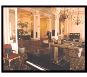 Elegant interior room with a grand piano in the foreground, ornate chandeliers hanging from the ceiling, patterned carpet, and classic upholstered furniture including sofas and chairs. Large mirrors and decorative columns enhance the spacious and luxurious ambiance.