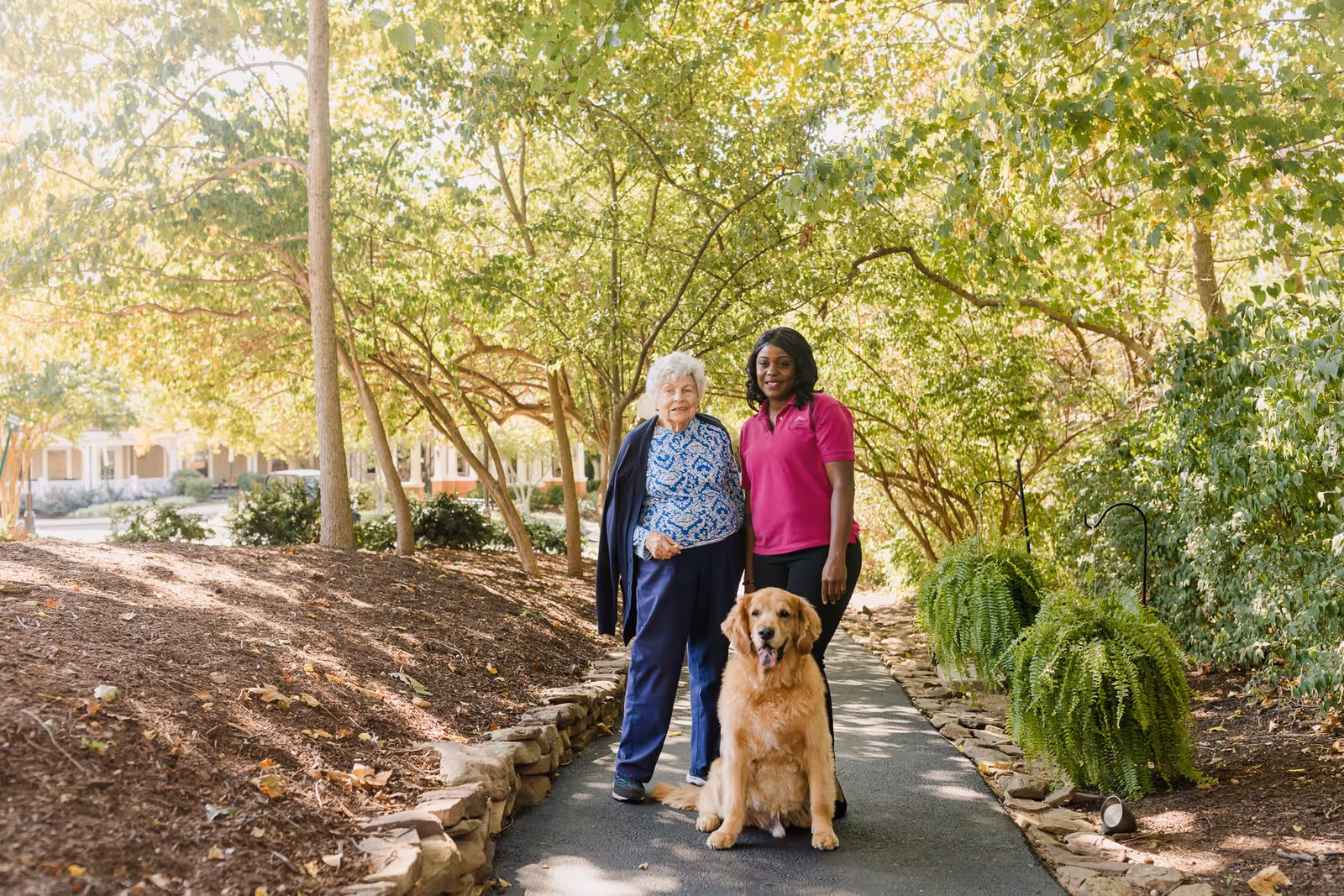 An elderly woman and a caregiver standing together on a paved garden path surrounded by trees and greenery, with a golden retriever sitting in front of them. The caregiver is wearing a pink shirt and the elderly woman is dressed in blue pants and a patterned top.