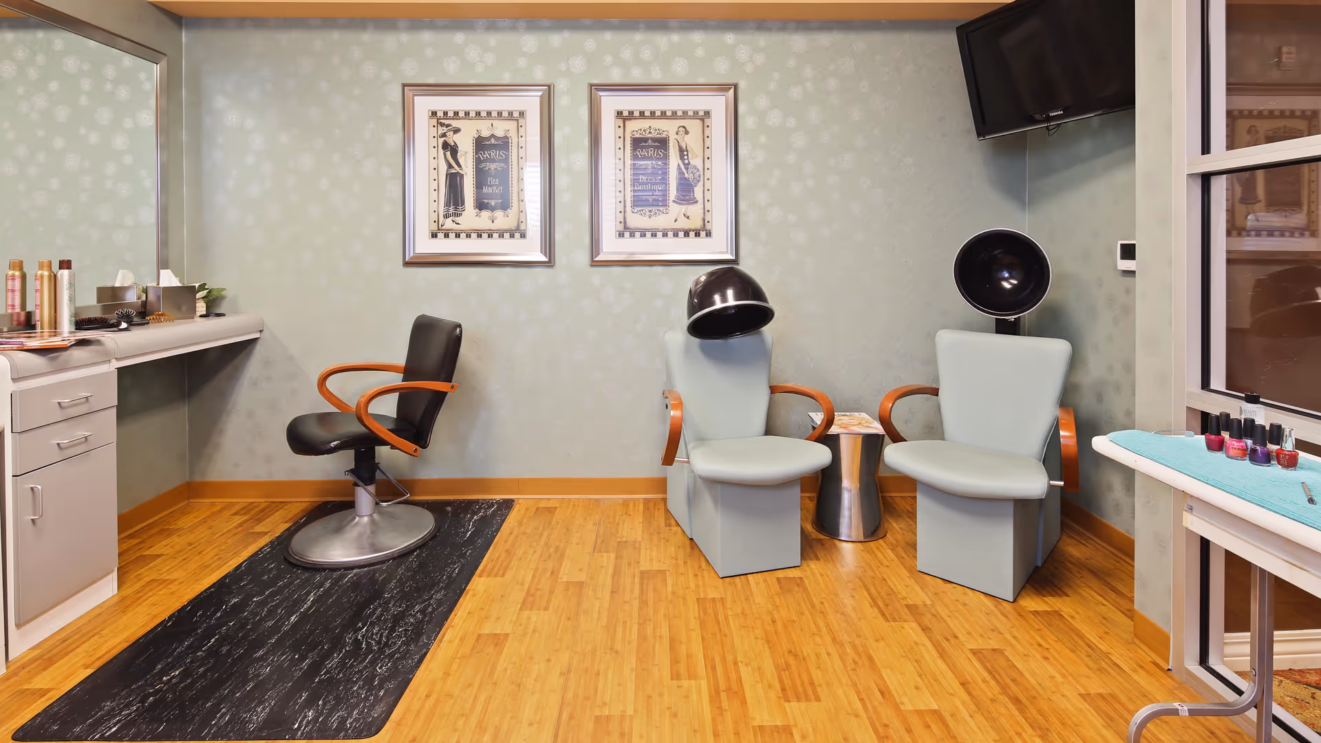 Interior of a salon area with a black salon chair on a black mat in front of a counter with hair products and tools. Two light blue chairs with wooden armrests are positioned next to each other, each with a black hair dryer hood above. Two framed vintage-style posters hang on the wall behind the chairs. A small metallic side table is placed between the two chairs. A TV is mounted on the wall in the corner, and a table with various nail polish bottles is visible near a window.