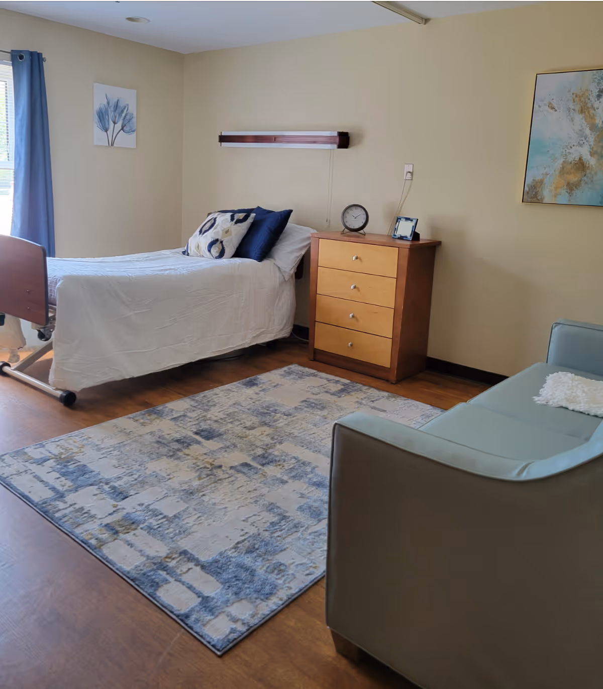 A neatly arranged bedroom in a senior living facility with a single bed covered in white bedding and decorative pillows. Next to the bed is a wooden chest of drawers with a clock and a small picture frame on top. A blue and beige patterned rug covers part of the wooden floor. There is a light blue armchair with a white cushion on it, and the walls are decorated with two pieces of artwork. A window with blue curtains lets in natural light.