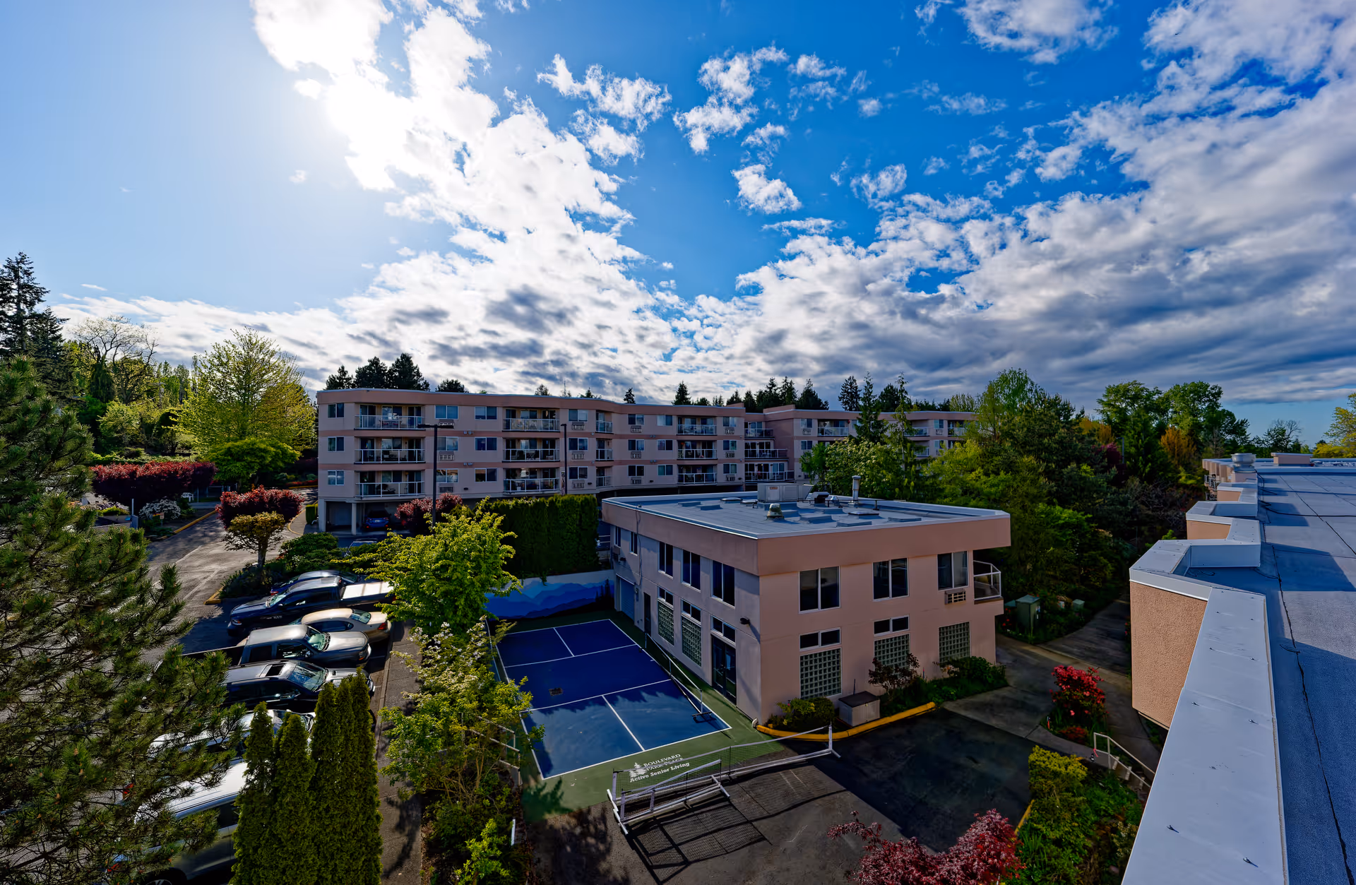 Aerial view of Boulevard Park Place Active Retirement Living facility showing multiple buildings surrounded by trees and greenery under a partly cloudy blue sky. There is a parking lot with cars and a small tennis or pickleball court visible between the buildings.
