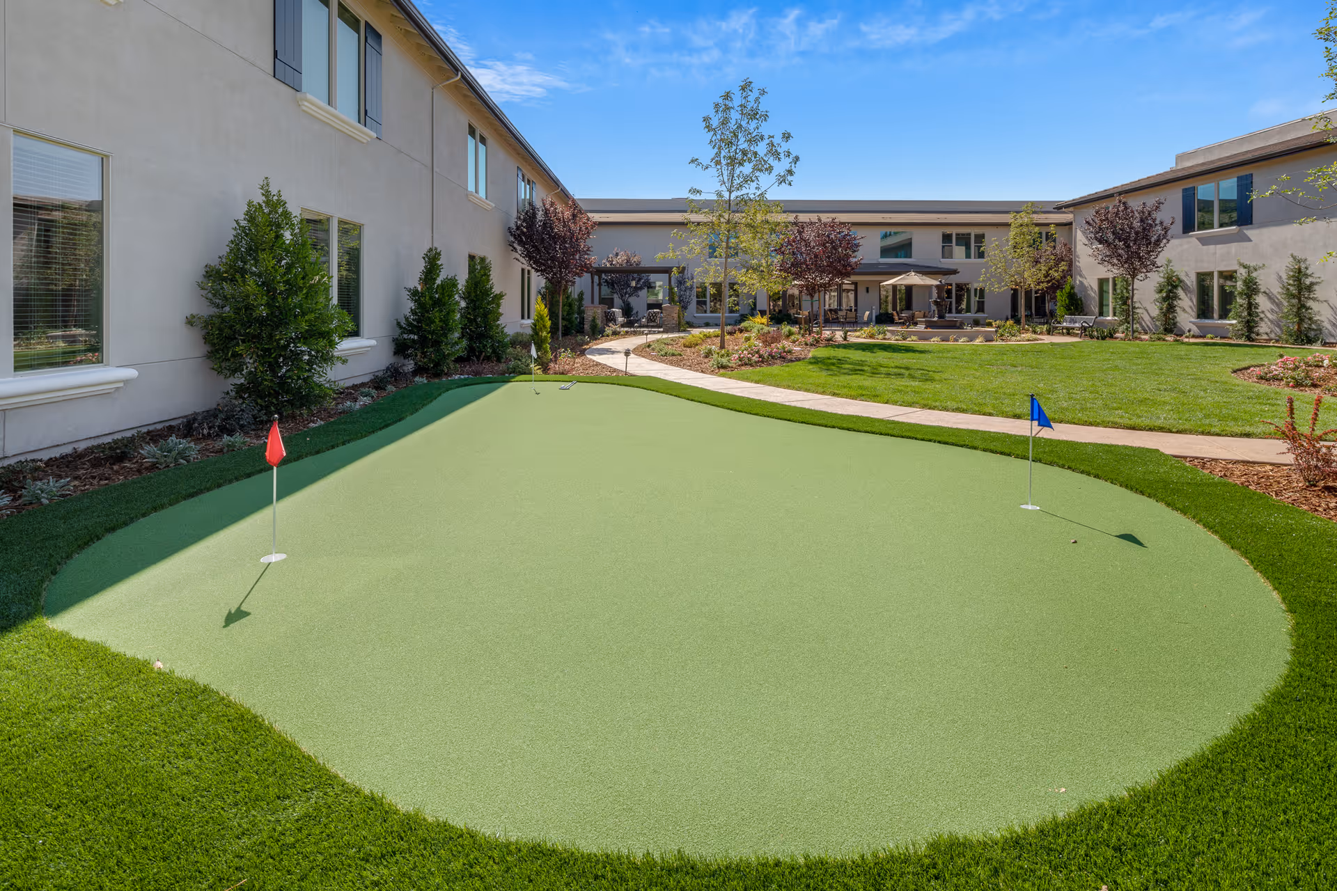Outdoor courtyard featuring a small putting green with red and blue flags in front of a two-story senior living building.