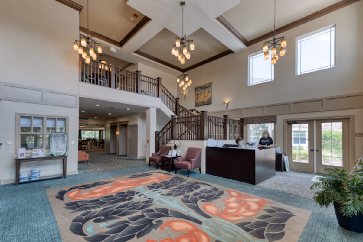 Spacious senior living facility lobby with a large decorative rug, two pink chairs with a small table, a reception desk with two staff members wearing masks, a staircase leading to an upper floor, large windows letting in natural light, and multiple hanging light fixtures.