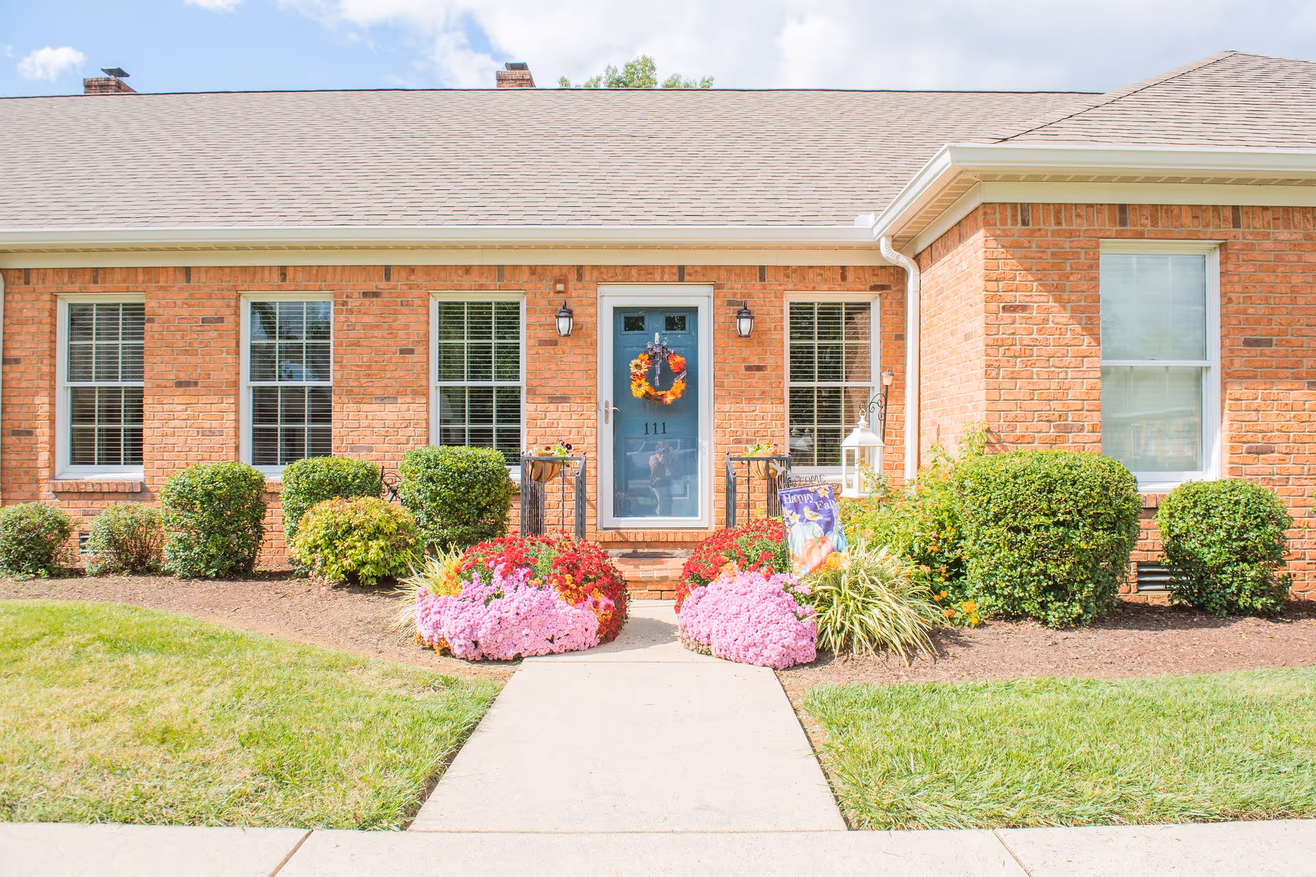 Front exterior view of a single-story brick building with a blue door decorated with a fall wreath. The entrance is flanked by two black lantern-style wall lights and surrounded by neatly trimmed bushes and colorful flower beds. A concrete walkway leads up to the door under a partly cloudy sky.