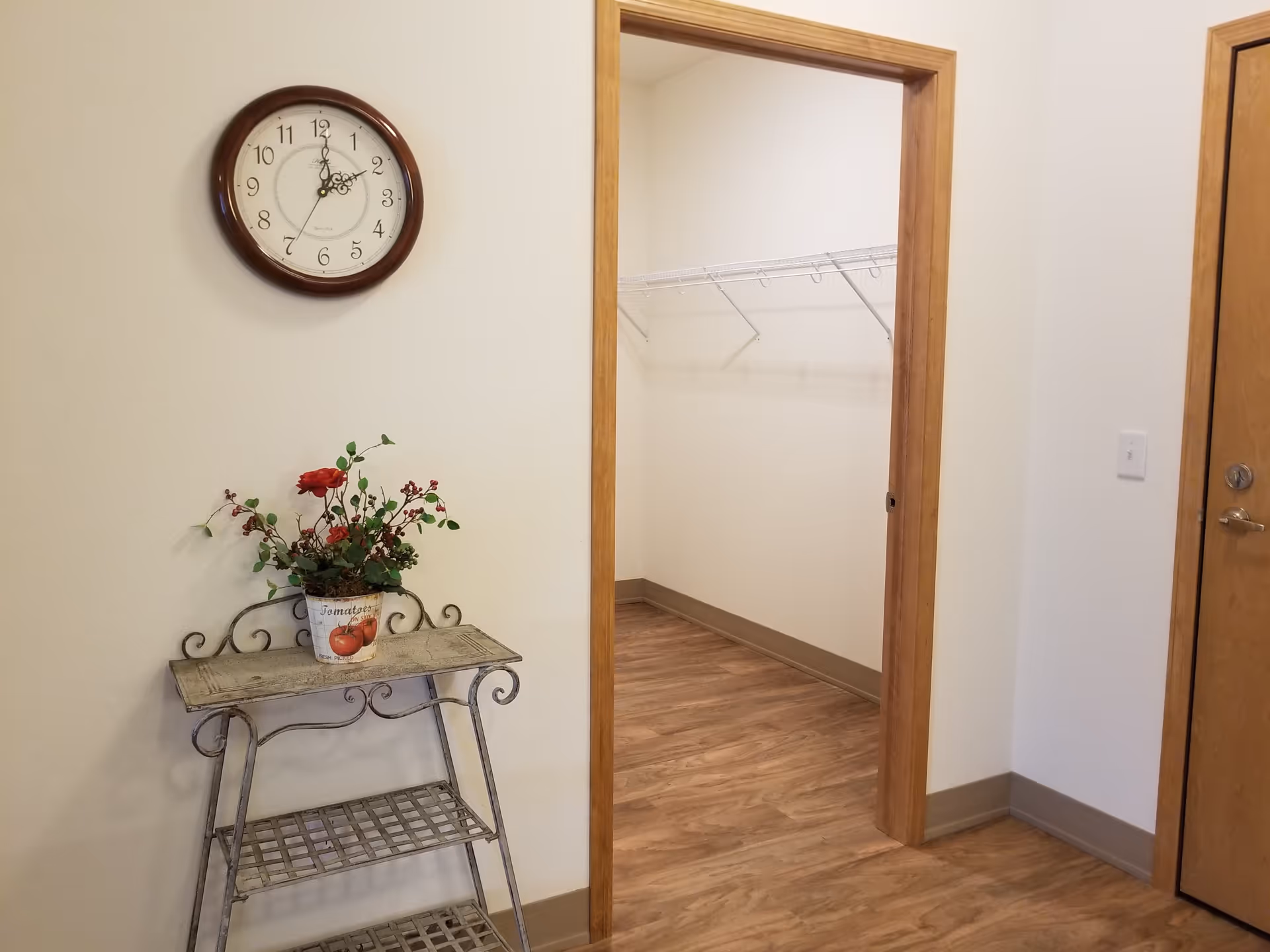 Interior view of a room with wooden flooring and white walls. A round wall clock with a dark brown frame is mounted on the wall showing the time as 1:35. Below the clock is a decorative metal shelf with a potted plant featuring red flowers and green leaves. To the right, there is an open doorway leading to a walk-in closet with white wire shelving. A closed wooden door with a silver handle is also visible on the right side of the image.