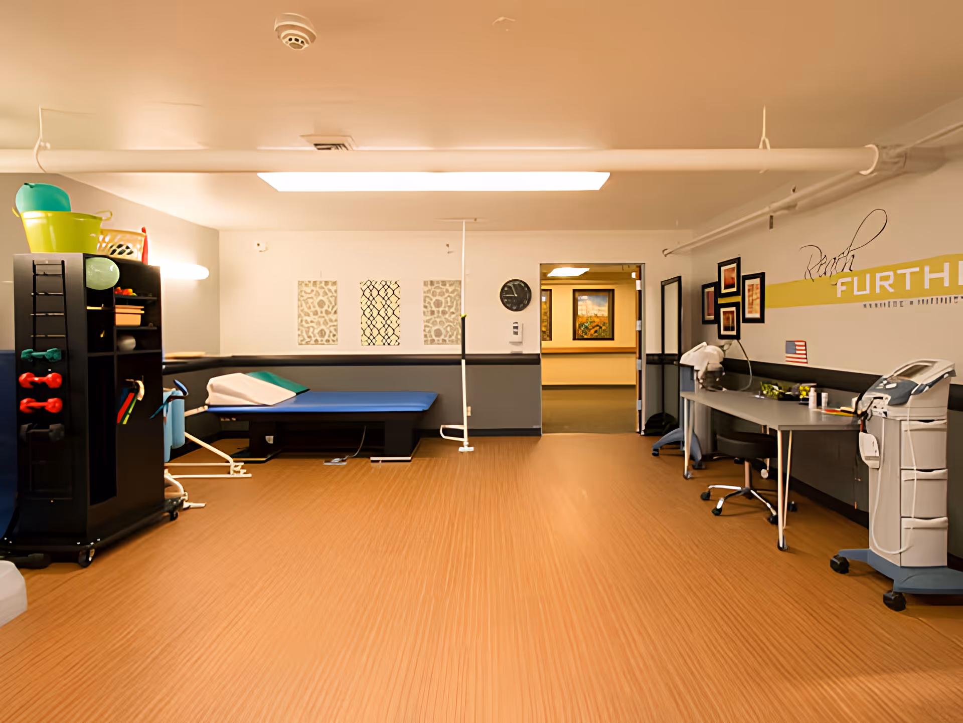 A physical therapy or rehabilitation room with a treatment table, exercise equipment including small dumbbells and resistance bands, a desk with a chair, medical equipment on a cart, and framed artwork on the walls. The room has wood-patterned flooring and neutral-colored walls with a yellow stripe and the word 'FURTH' partially visible.