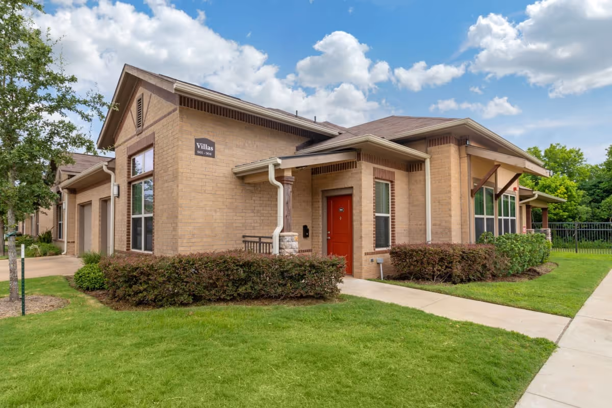 Exterior view of a single-story brick villa building with a red door, large windows, and a small covered porch. The building is surrounded by green grass, shrubs, and a concrete walkway under a partly cloudy blue sky.