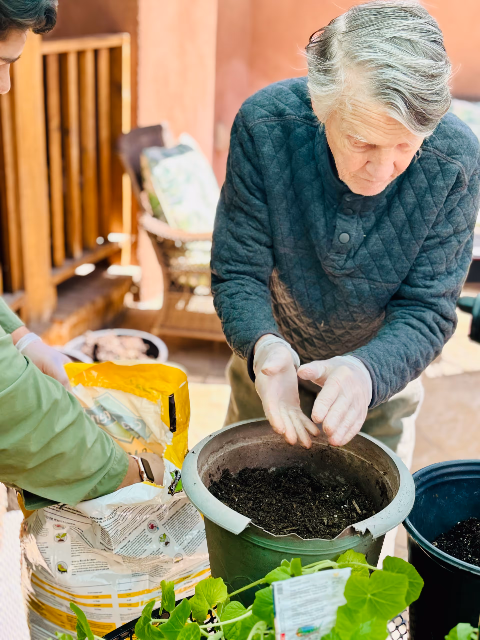 An elderly person wearing a dark quilted jacket and gloves is planting seeds or working with soil in a large green pot outdoors. Another person in a green jacket is assisting by handling a bag of soil or potting mix. There are green plants and gardening supplies on a table nearby, with a wooden fence and outdoor furniture in the background.