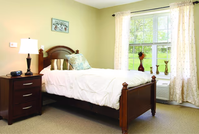 Bright bedroom with a wooden single bed, nightstand and lamp, and a window with curtains overlooking a green lawn.