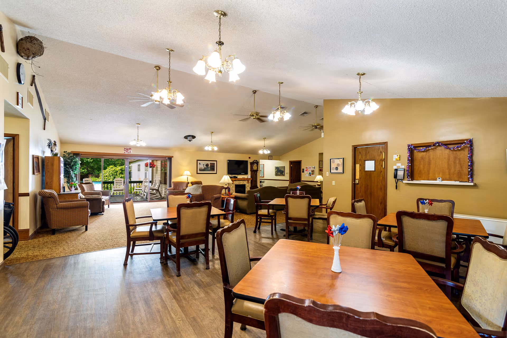 A spacious senior living common area with multiple wooden tables and cushioned chairs arranged for dining or socializing. The room features warm beige walls, several ceiling light fixtures, ceiling fans, and a carpeted seating area with armchairs near large sliding glass doors that open to an outdoor patio with greenery. There is a fireplace with a TV mounted above it and various framed pictures on the walls.