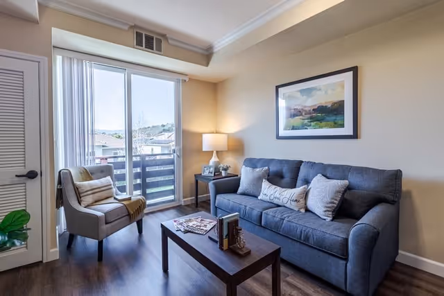 A cozy living room with a dark gray sofa adorned with three pillows, a wooden coffee table with books and magazines, a side table with a lamp and framed photo, a beige armchair with a pillow and throw blanket, and a large window with vertical blinds letting in natural light.