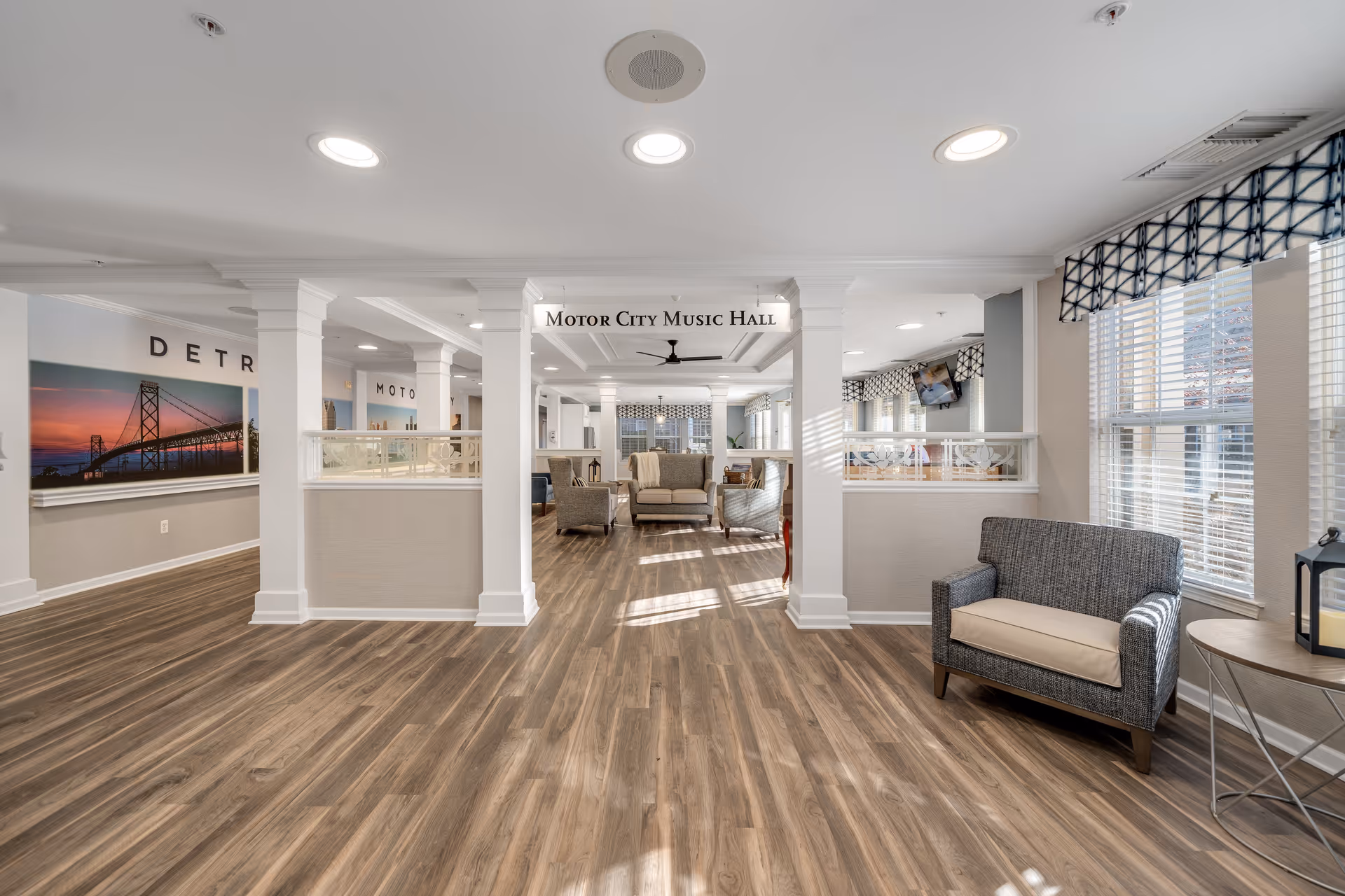A spacious and well-lit common area with wood flooring and white walls. The room features several seating arrangements including cushioned chairs and sofas. Large windows with patterned valances allow natural light to fill the space. A sign hanging from the ceiling reads 'Motor City Music Hall'.