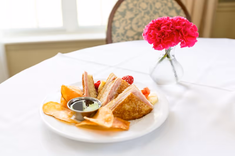 A plate with a sandwich cut into triangles, potato chips, a small container of dipping sauce, and some fruit pieces on a white tablecloth. A small vase with bright pink flowers is placed next to the plate on the table.