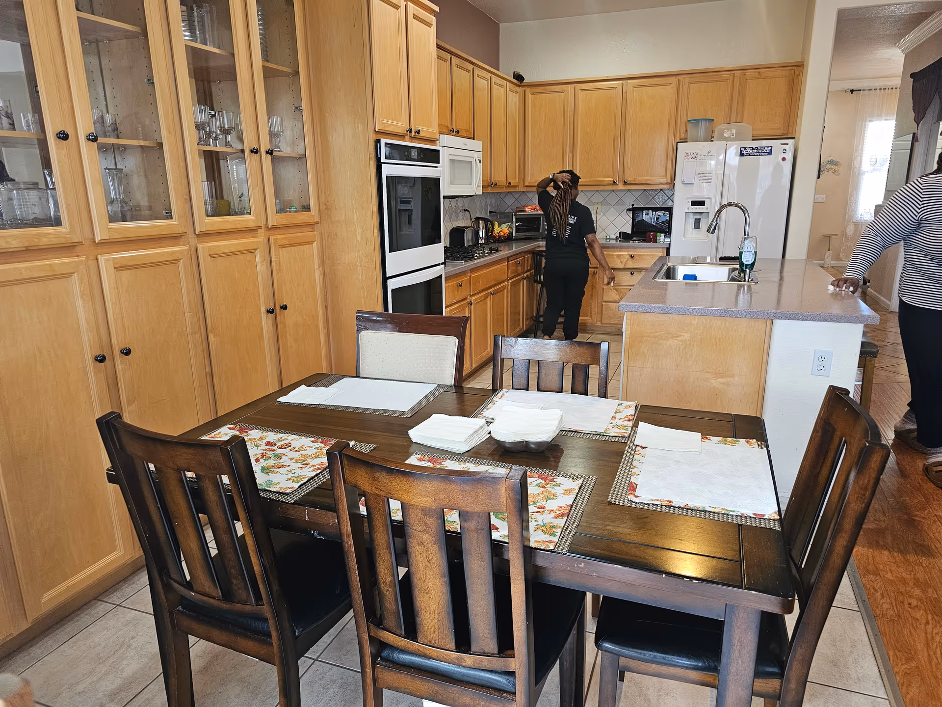 A kitchen and dining area with wooden cabinets and a dark wooden dining table set with six chairs. The table has placemats and napkins arranged on it. In the background, a person is standing near the kitchen counter and another person is partially visible on the right side. The kitchen features a white refrigerator, built-in oven, microwave, and a sink on the island counter.