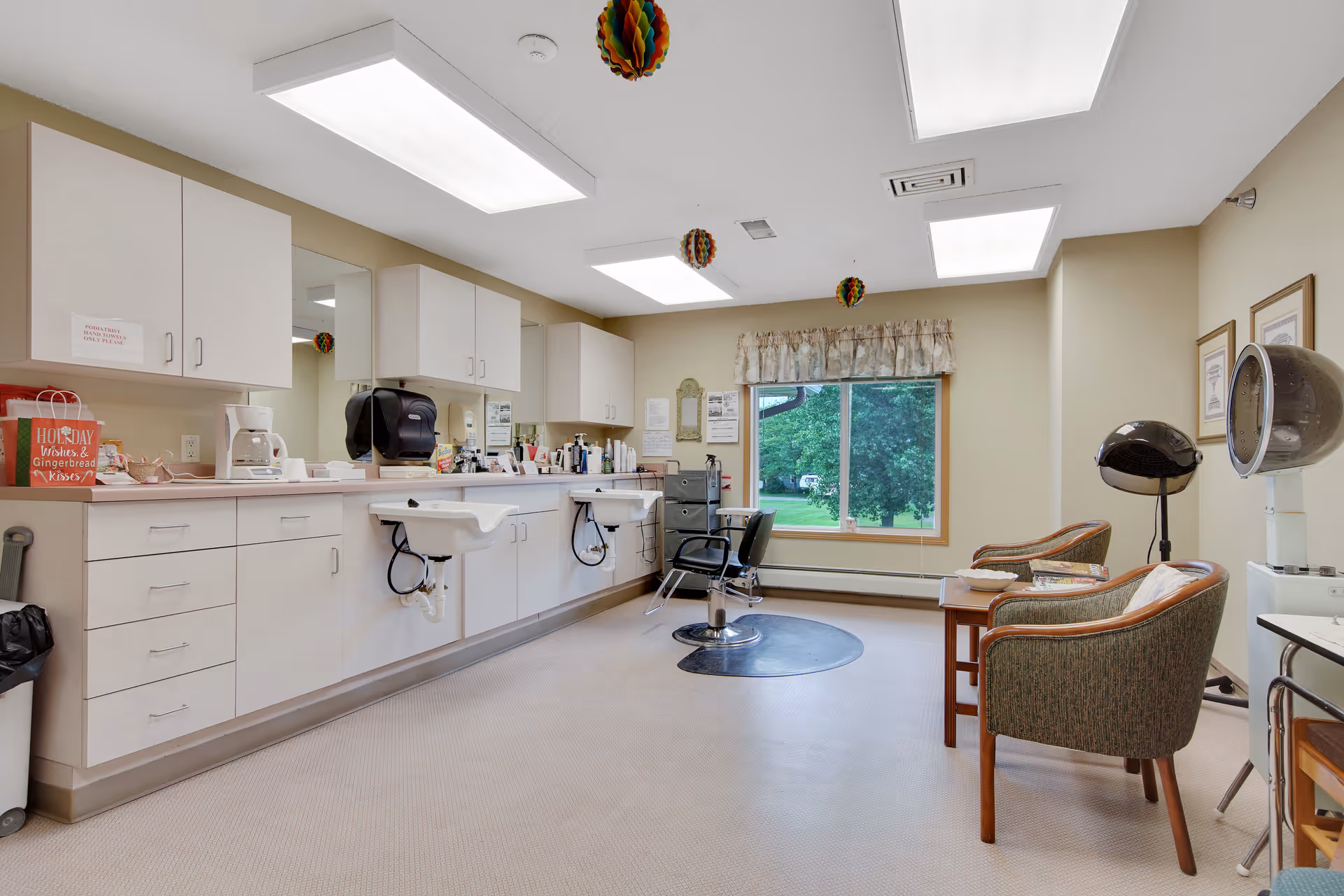 A senior living salon with styling chairs, shampoo sinks, hooded hair dryers, and seating by a large window.