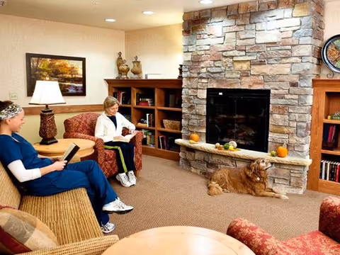 A cozy living room with two people sitting and reading. One person is dressed in blue scrubs and the other in casual clothes. There is a stone fireplace with small pumpkins and decorative items on the mantel. A golden retriever dog is lying on the carpet in front of the fireplace. The room has wooden bookshelves, a table lamp, and comfortable upholstered chairs and sofa.