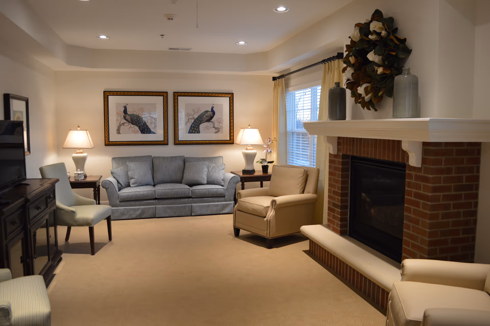 A cozy living room with a blue sofa, two beige armchairs, and two side tables with lamps. There is a brick fireplace decorated with a wreath and vases, and two framed peacock pictures hang on the wall above the sofa. A window with cream-colored curtains lets in natural light.