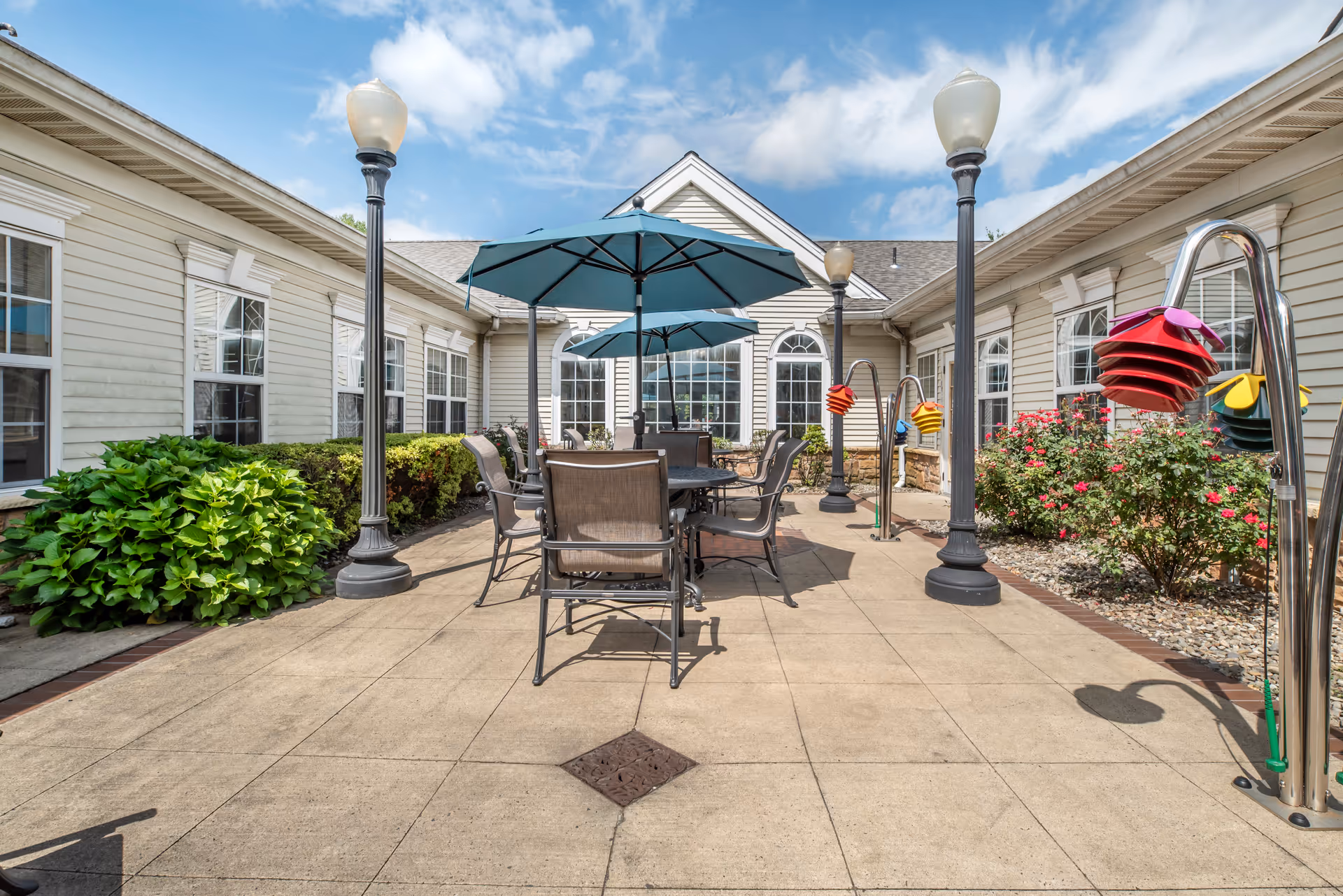 Outdoor patio area at Brookdale Hamilton with tables and chairs under blue umbrellas, surrounded by beige building walls with windows, lamp posts, green bushes, and colorful hanging bird feeders.