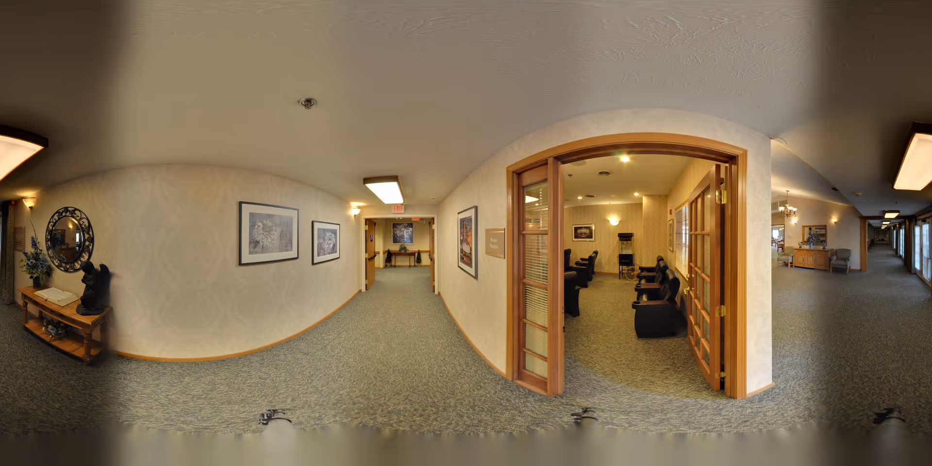 A wide hallway in a senior living facility with carpeted floors and beige patterned wallpaper. On the left side, there is a wooden table with decorative items including a mirror and a sculpture. The hallway leads to a room with glass-paneled double doors, revealing several black chairs arranged inside. The hallway is well-lit with ceiling lights and wall sconces, and framed artwork is displayed on the walls.