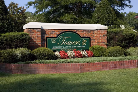 A brick sign surrounded by neatly trimmed bushes and flowers, displaying the name 'The Towers Retirement & Assisted Living Community' with an address below it, set against a backdrop of trees and greenery.