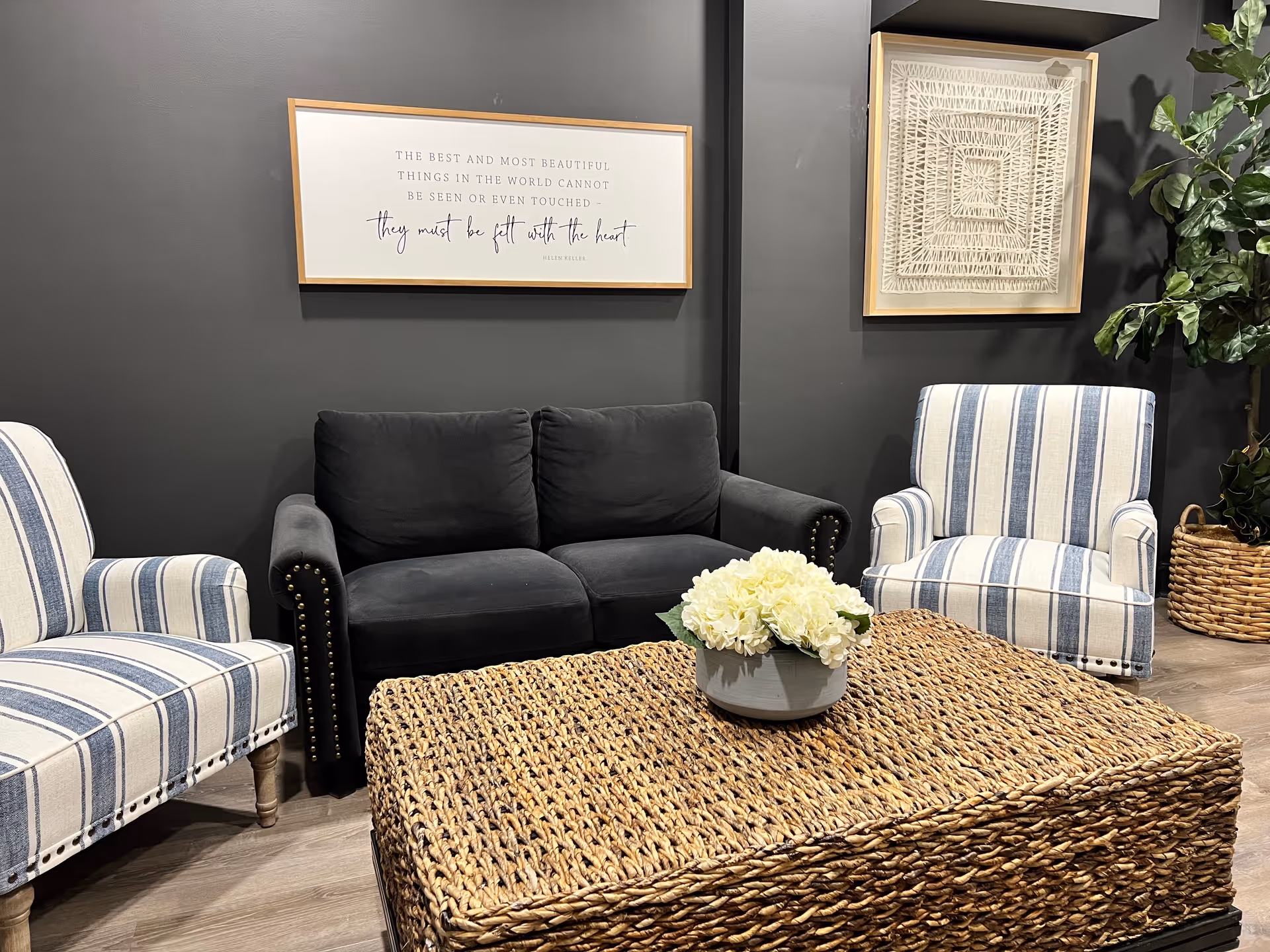 A cozy living room area with a dark gray couch flanked by two striped armchairs. A woven rattan coffee table with a white flower arrangement sits in front of the seating. The walls are dark gray and decorated with two framed pieces of art, one featuring a quote by Helen Keller. A green potted plant is visible in the corner.
