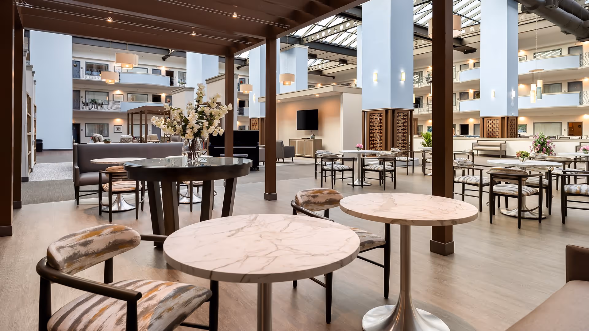 Spacious and modern common area in a senior living facility with multiple round marble tables and cushioned chairs arranged under a wooden pergola structure. The area is well-lit with natural light coming from a large glass ceiling, and there are additional seating areas and a TV in the background. The space has a clean, open design with light wood flooring and white walls accented by wooden columns.