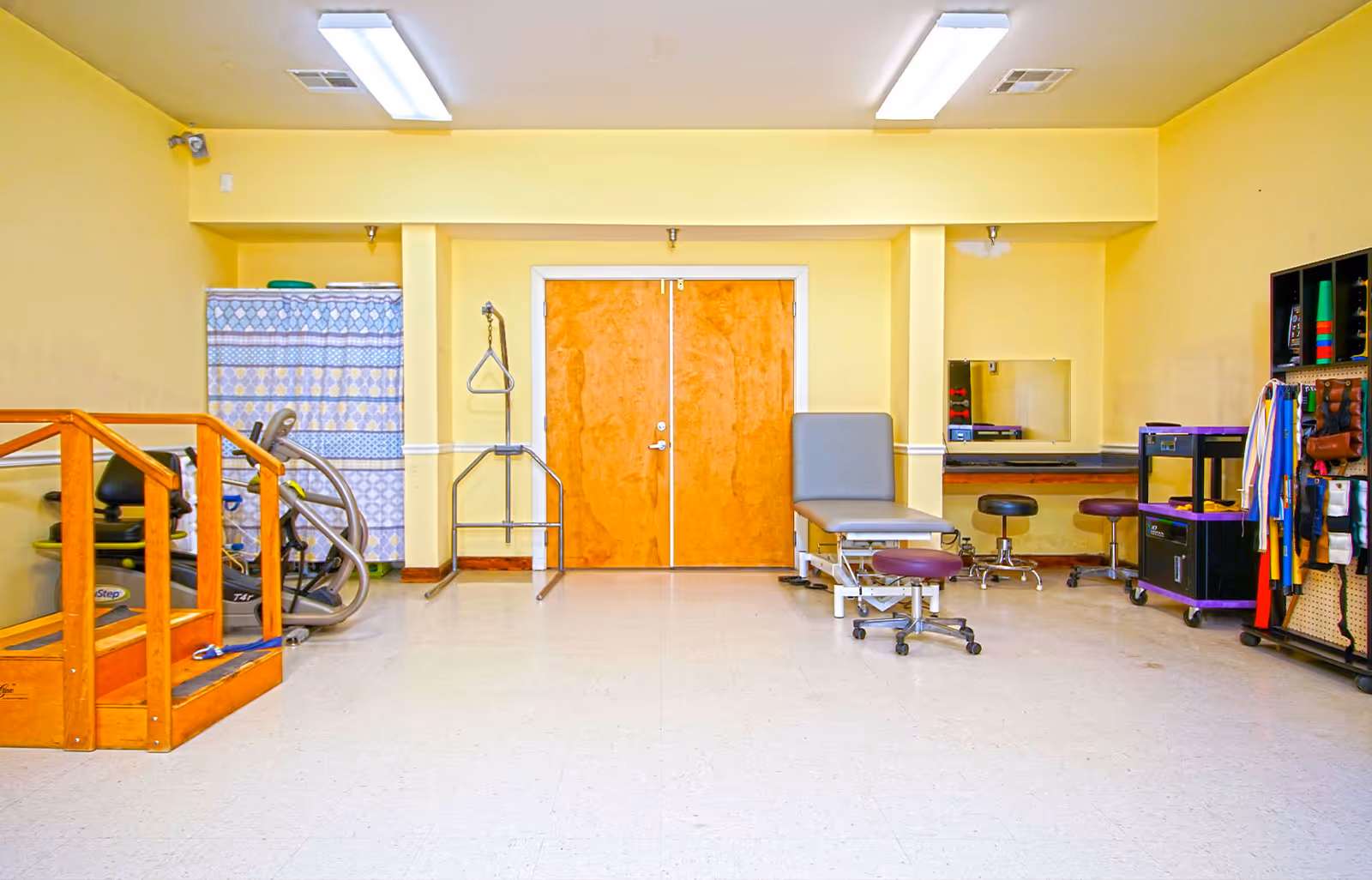 A rehabilitation or physical therapy room with yellow walls and a white tiled floor. The room contains exercise equipment including a wooden staircase for step exercises, an exercise bike, a therapy table with stools, a mirror on the wall, and various therapy tools and supplies organized on shelves and racks. There is a double wooden door in the center of the back wall.