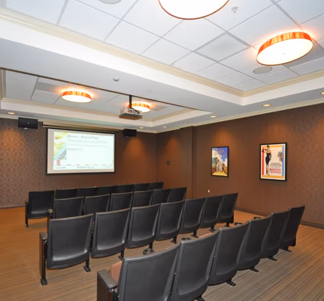 A small theater or presentation room with rows of black chairs facing a projection screen. The screen displays information about a movie word play event. The room has brown walls with two framed posters and ceiling lights.