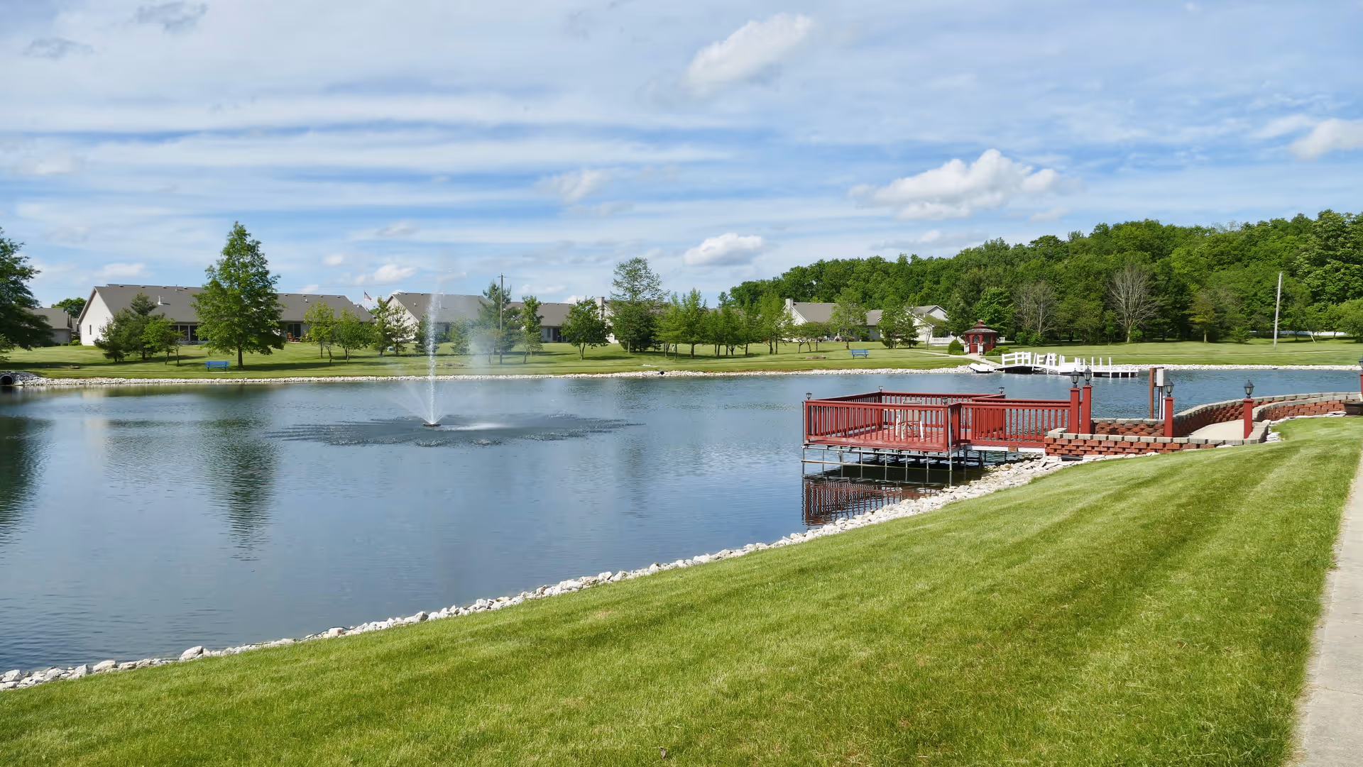 A peaceful outdoor scene at The Cedars featuring a pond with a water fountain in the center, surrounded by green grass and trees. There is a red wooden dock extending into the pond, and residential buildings are visible in the background under a partly cloudy sky.