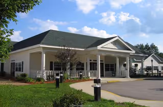 Exterior view of a single-story building with a covered entrance and white columns, surrounded by green grass and trees under a partly cloudy sky.