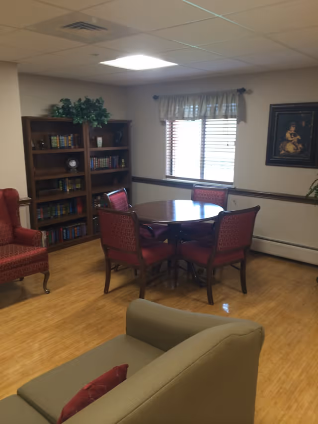 A cozy common room with a round wooden table surrounded by red-upholstered chairs, bookshelves, armchairs, and a window with blinds.