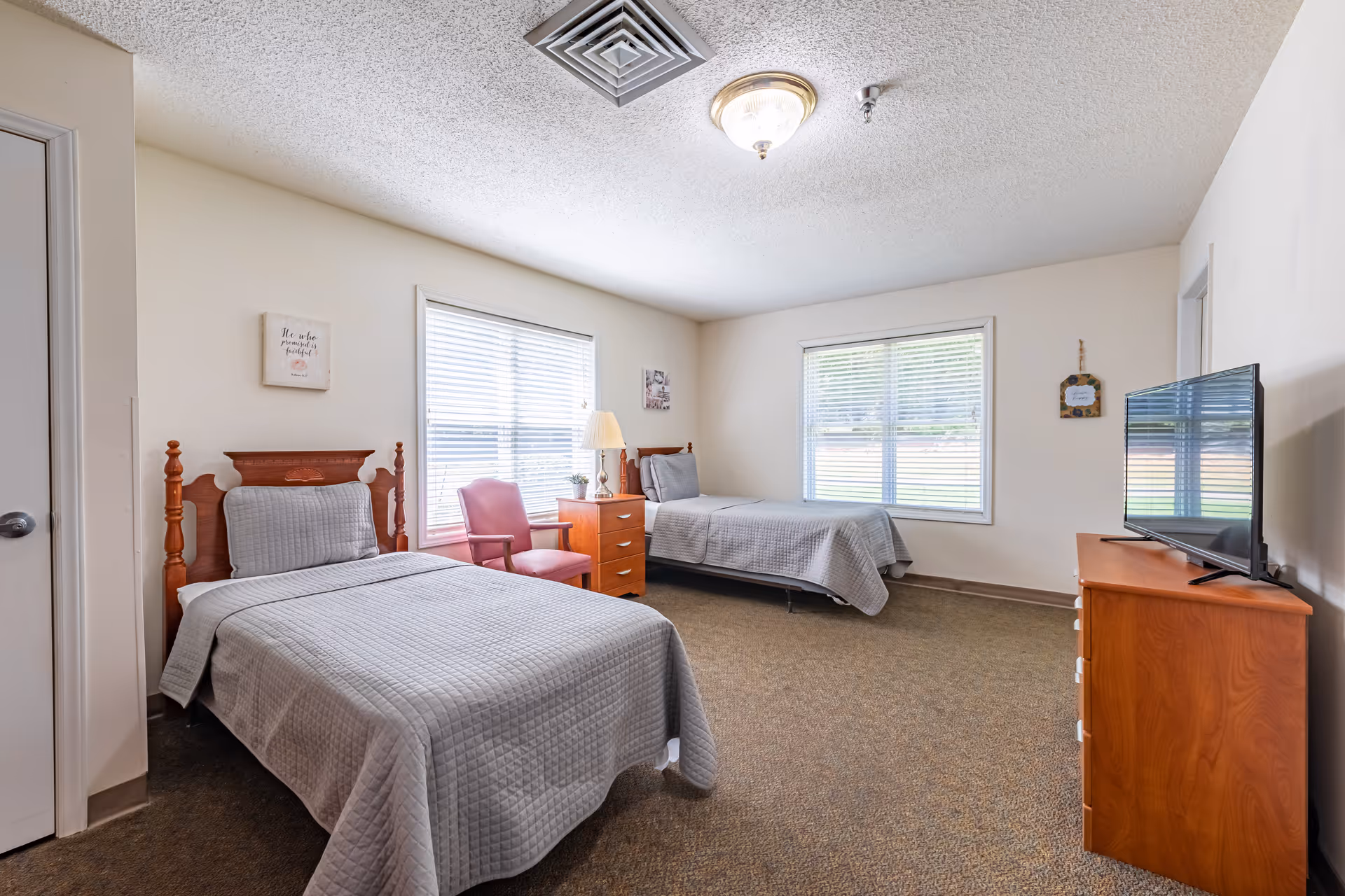 A bright bedroom with two single beds covered in gray quilted bedspreads. Between the beds is a wooden nightstand with a lamp and a small plant. A pink upholstered chair is placed next to the nightstand. The room has two large windows with white blinds, beige walls, and a carpeted floor. A wooden dresser with a flat-screen TV on top is positioned against the wall opposite the beds. The ceiling has a light fixture and an air vent.