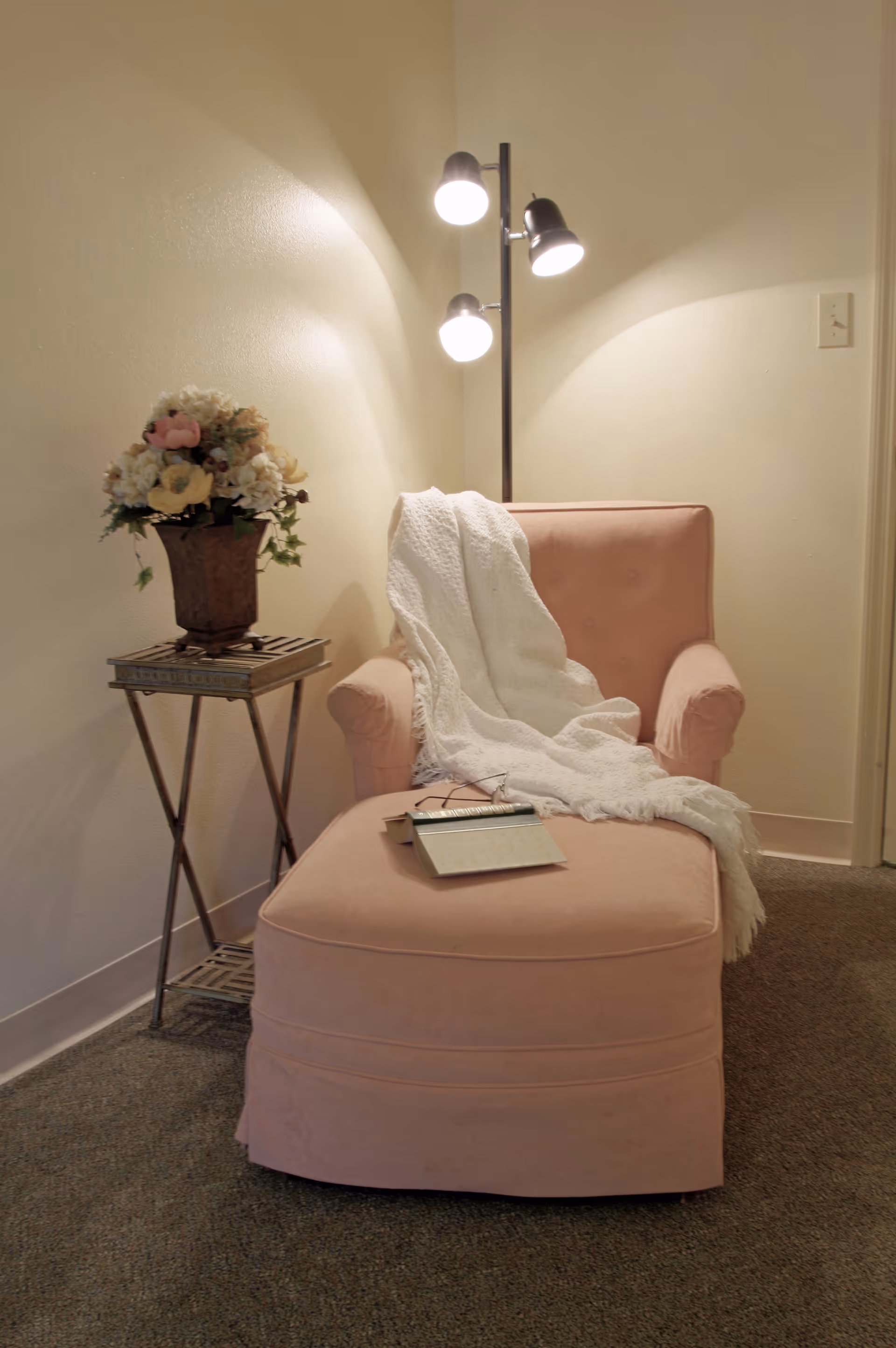 A cozy corner with a pink upholstered armchair and matching ottoman. A white knitted throw blanket is draped over the chair, and a book with reading glasses rests on the ottoman. Next to the chair is a small metal side table holding a decorative flower arrangement. A floor lamp with three lights illuminates the area against a plain beige wall.