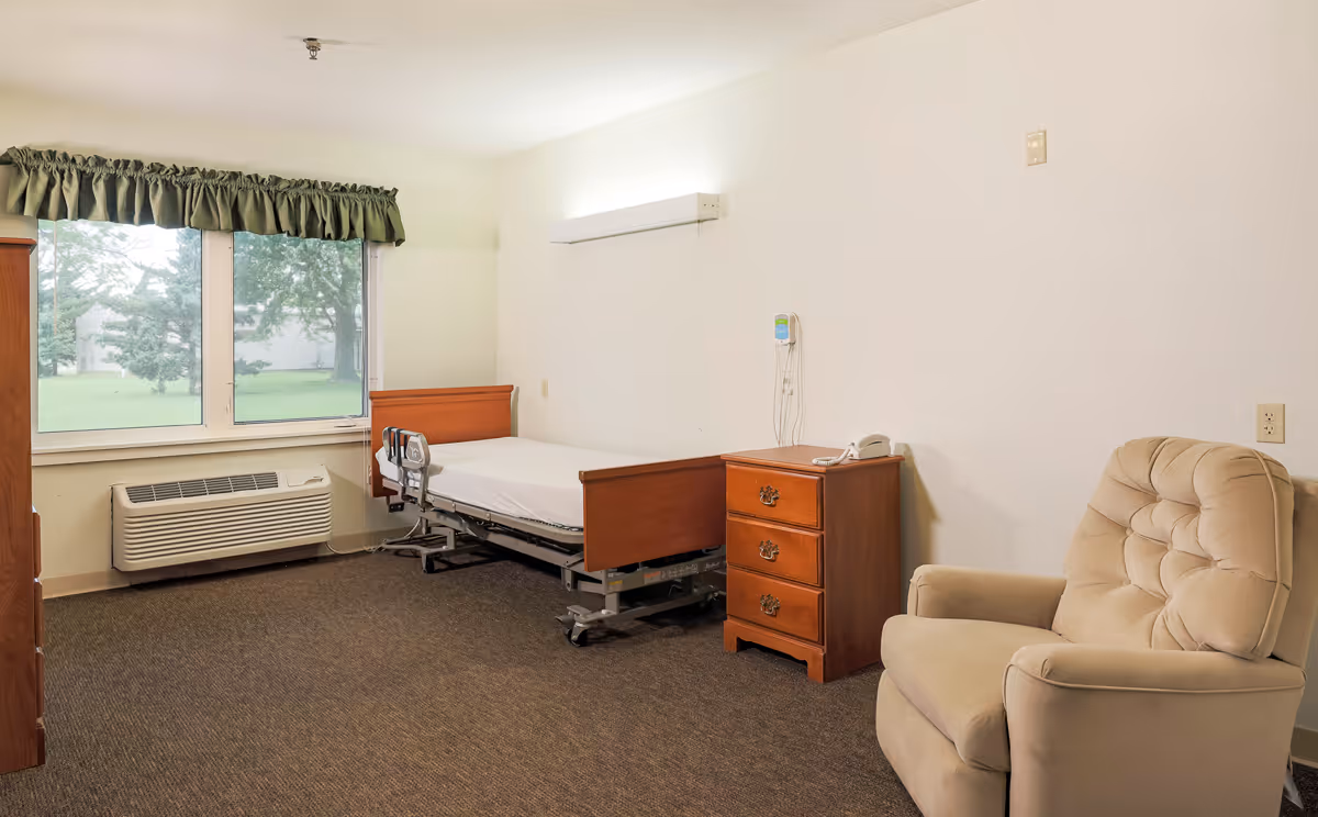 A simple nursing-home bedroom with a hospital bed, wooden bedside dresser, beige recliner, and a large window with a green valance.