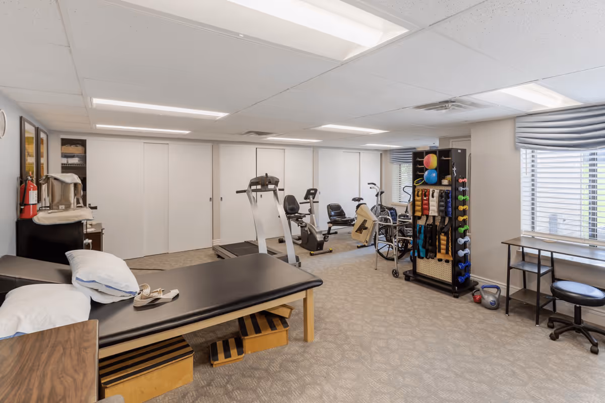 A senior living facility exercise and therapy room with a black padded therapy table with pillows, exercise equipment including stationary bikes and a treadmill, a rack with colorful exercise balls, dumbbells, and resistance bands, a wheelchair, and a desk with a rolling chair near a window with blinds and a blue valance.