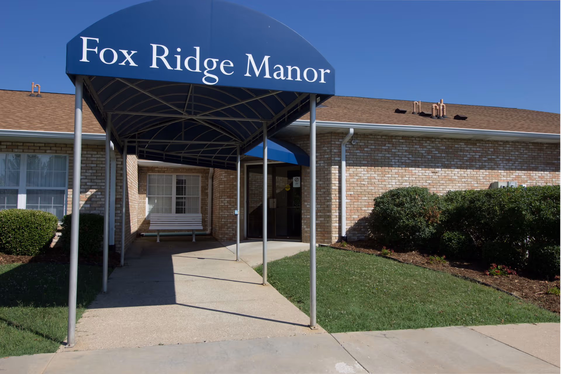 Entrance of Fox Ridge Manor with a blue canopy over a sidewalk leading to the front door of a single-story brick building.