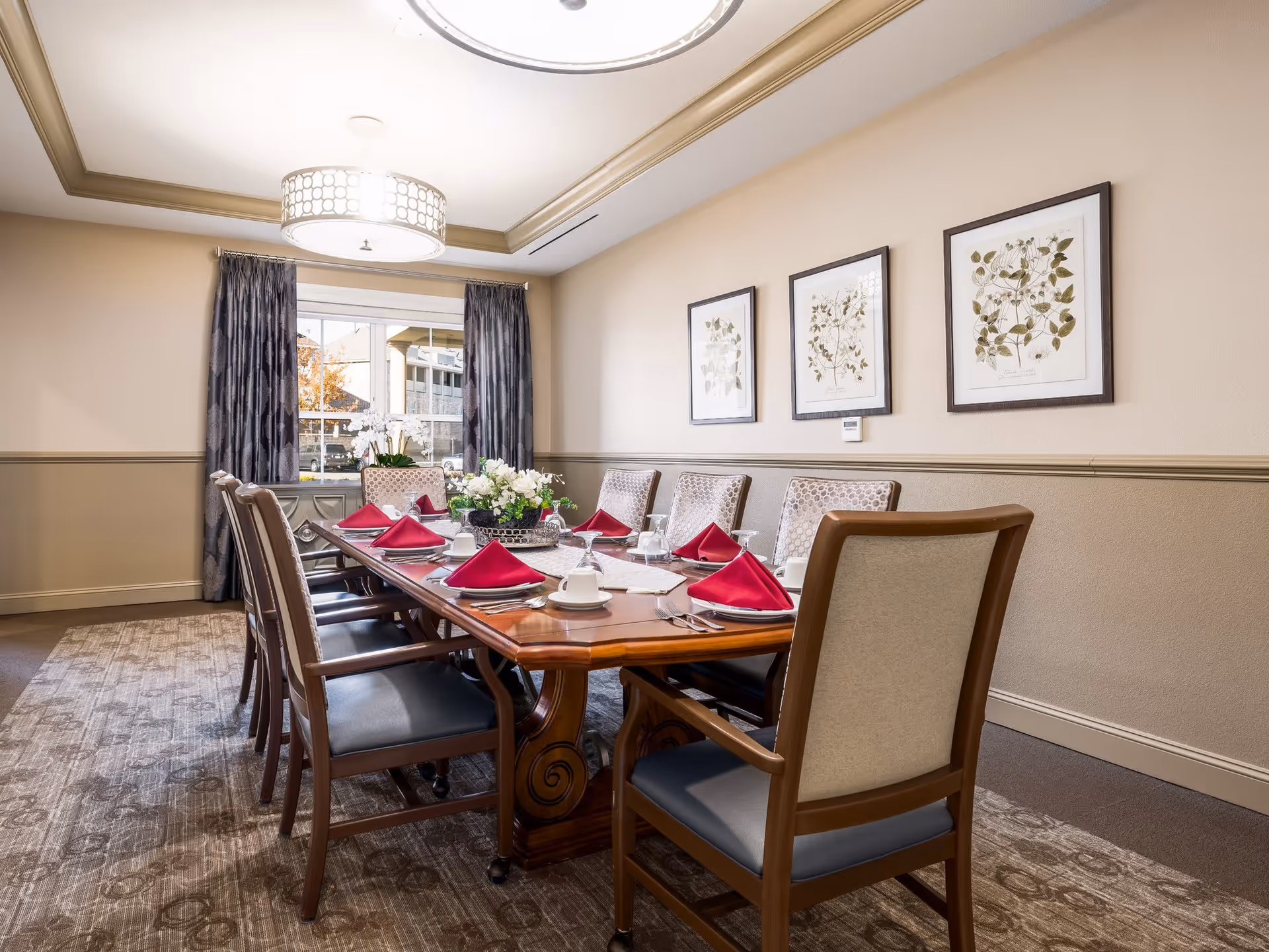 A formal dining room with a long wooden table set for eight people. Each place setting has a white cup and saucer, silverware, a wine glass, and a red folded napkin. The room has beige walls with three framed botanical prints, a window with dark curtains, a patterned carpet, and a modern ceiling light fixture.