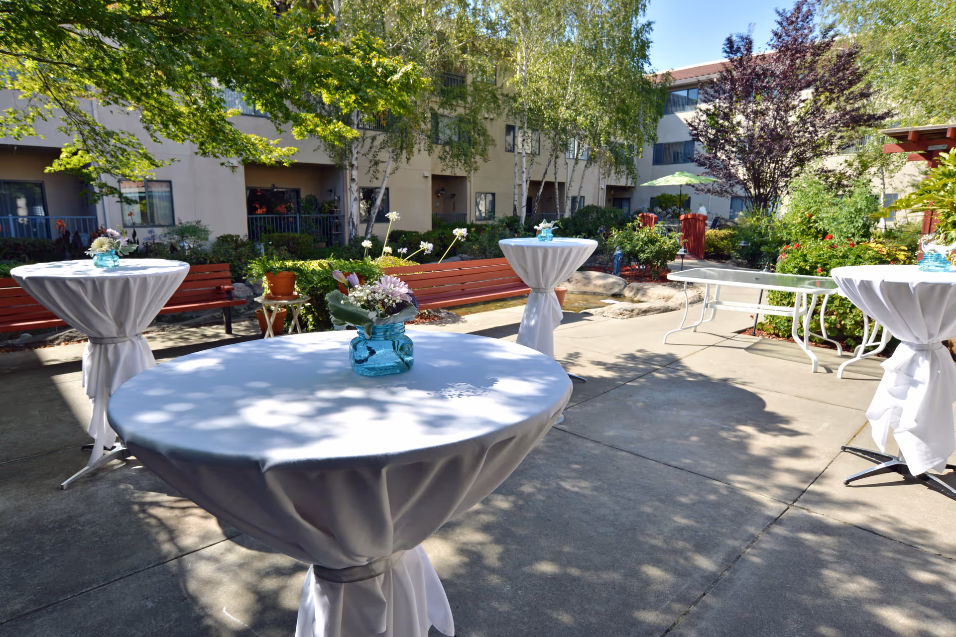 Outdoor courtyard area with round cocktail tables covered in white tablecloths and decorated with small flower arrangements in blue vases. There are red benches, green bushes, trees, and a multi-story building in the background under a sunny sky.