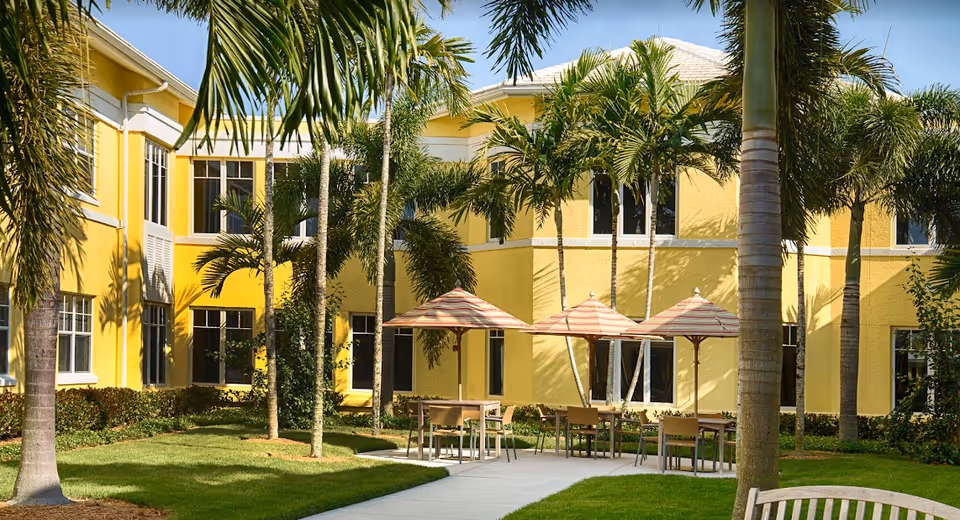 Outdoor courtyard area of a yellow two-story building with several palm trees and green grass. There are tables with striped umbrellas and chairs arranged on a paved area, creating a relaxing outdoor seating space.