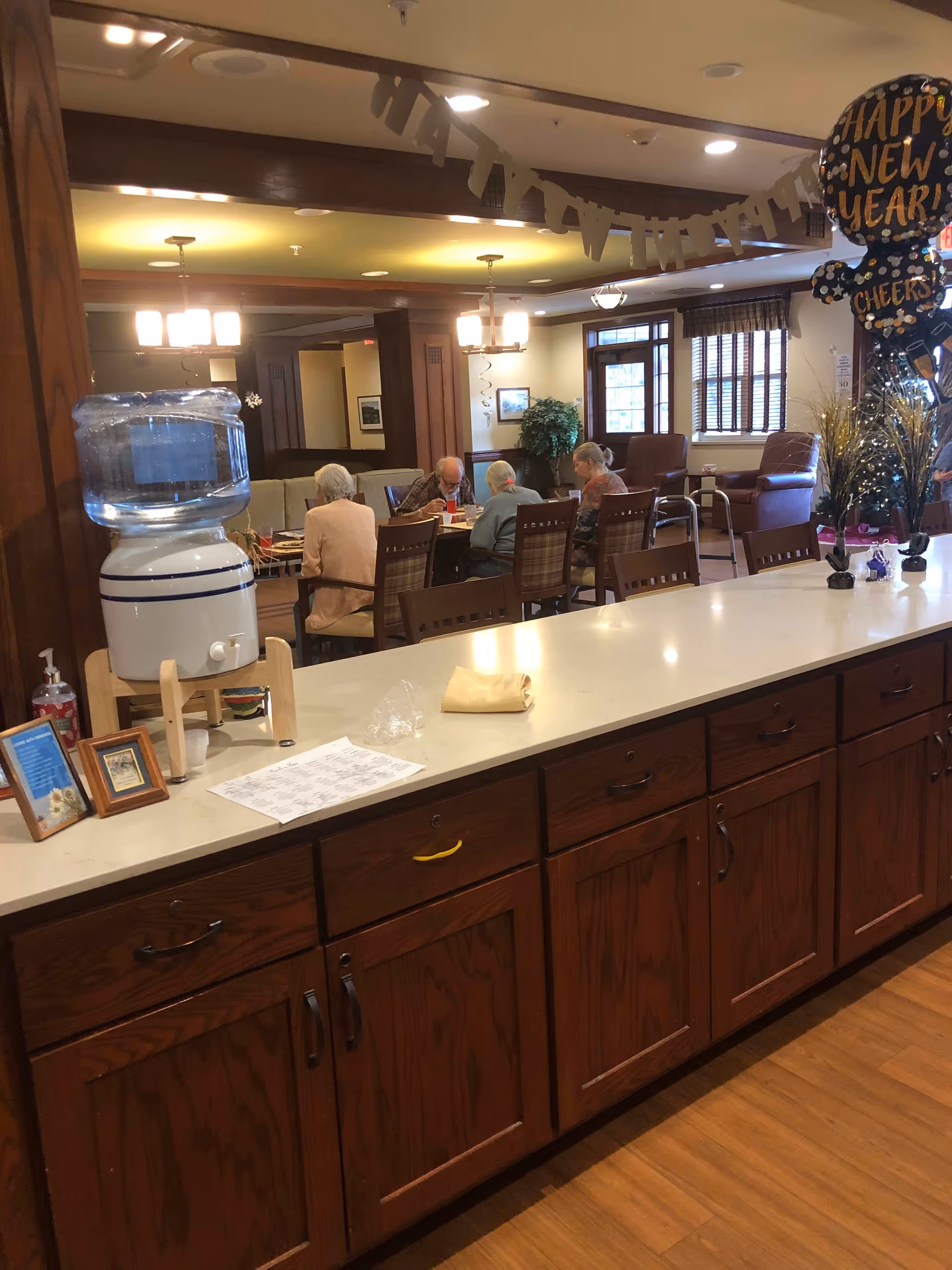 Interior view of a senior living facility common area with a long wooden counter in the foreground. On the counter, there is a water dispenser, a few framed pictures, a paper, and some small decorative items. In the background, four elderly people are seated around a table engaged in an activity. The room is warmly lit with ceiling lights and decorated with a 'Happy New Year' balloon and hanging decorations.