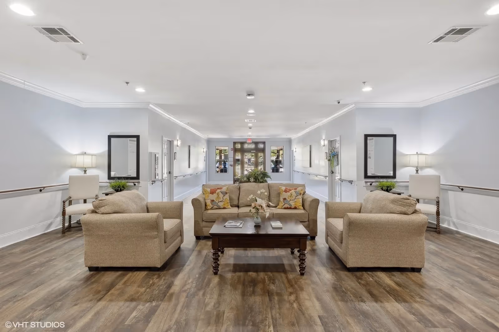A spacious and well-lit common area in a senior living facility featuring a beige sofa and two matching armchairs arranged around a dark wooden coffee table with decorative items. The room has light blue walls, wooden flooring, and two side tables with lamps and plants. There are handrails along the walls and a glass door at the far end of the hallway.