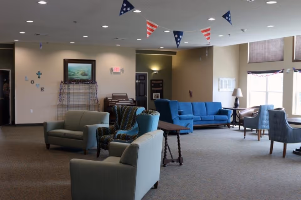 A spacious living room area with various seating options including blue and beige armchairs, a blue sofa, and a small table with a lamp near large windows. The ceiling is decorated with red, white, and blue triangular pennant banners. On the far wall, there is a framed painting, a small organ, and decorative letters spelling 'LOVE'.