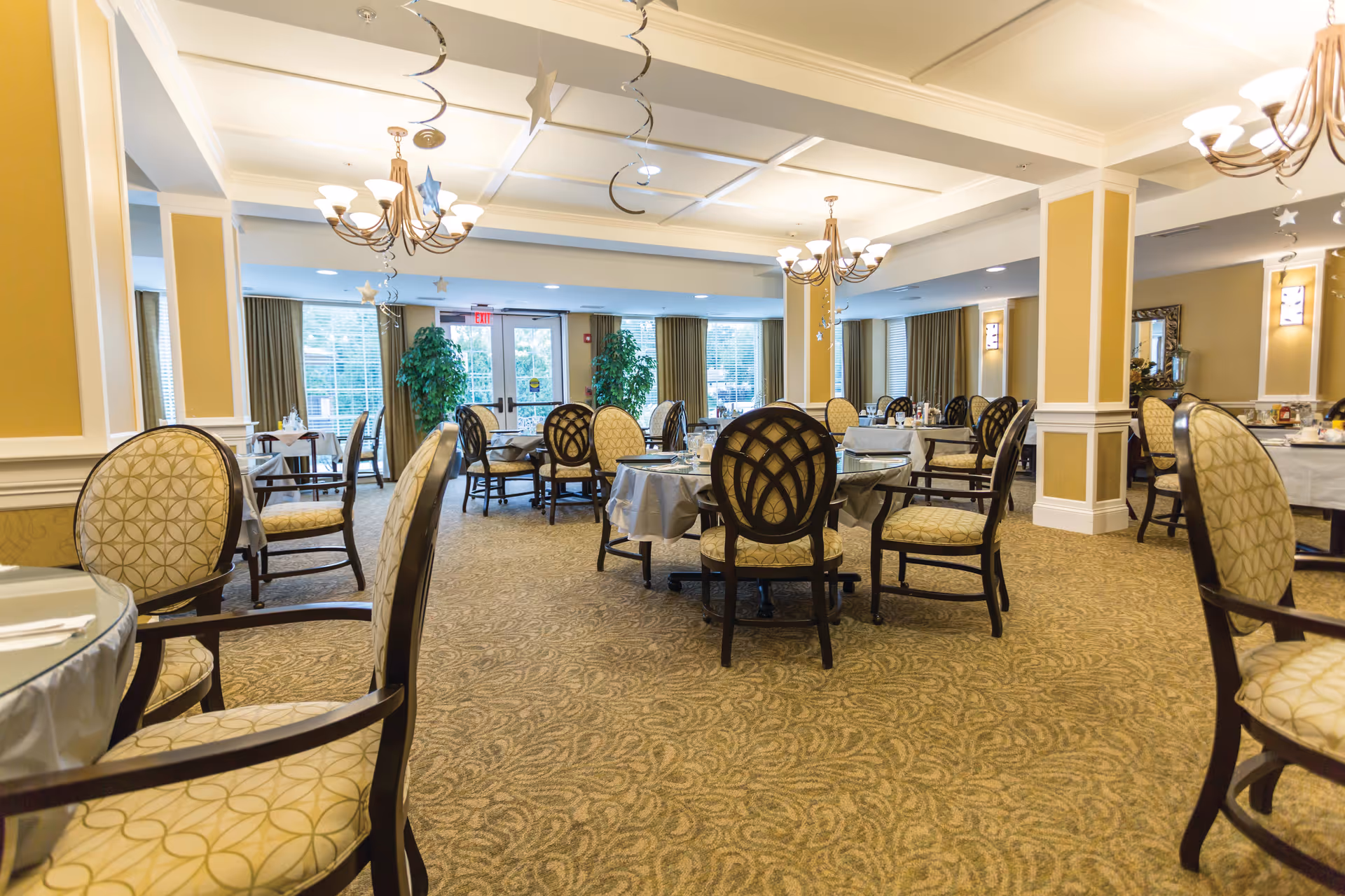 A spacious dining room in an assisted living facility with round tables covered with white tablecloths and surrounded by cushioned chairs with patterned upholstery. The room has beige walls with white trim, carpeted floors, chandeliers hanging from the ceiling, and large windows letting in natural light. Decorative plants and curtains are visible near the windows.