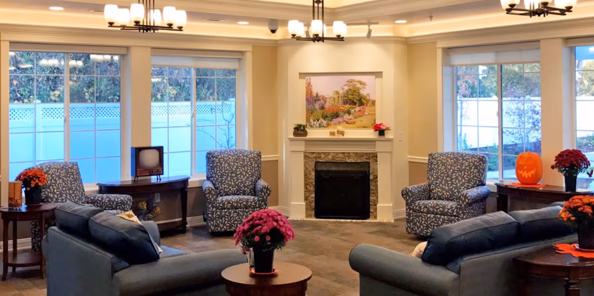A cozy living room with two blue patterned armchairs and two gray sofas arranged around a fireplace. The fireplace has a floral painting above it and small decorative items on the mantel. Large windows with white blinds let in natural light, and there are several potted flowers and a carved pumpkin on side tables. The ceiling has recessed lighting and two chandeliers.