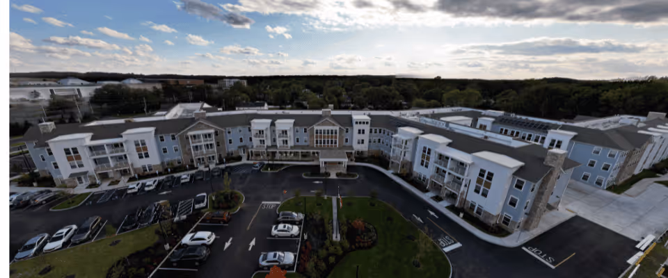 Aerial front view of a large multi-story senior living facility with parking lot and landscaped entrance under a partly cloudy sky.